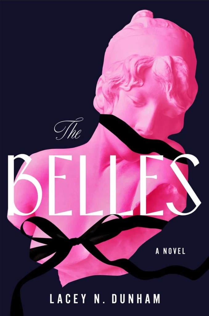 Book cover for The Belles: A Novel by Lacey N. Dunham. The design features a bright pink classical-style bust of a woman against a dark navy background. A black ribbon wraps around the statue’s neck and across its mouth, partially covering it. The title “The Belles” appears in large white letters, with “The” in elegant script and “A Novel” and the author’s name below in smaller text. The overall tone is striking and dramatic.